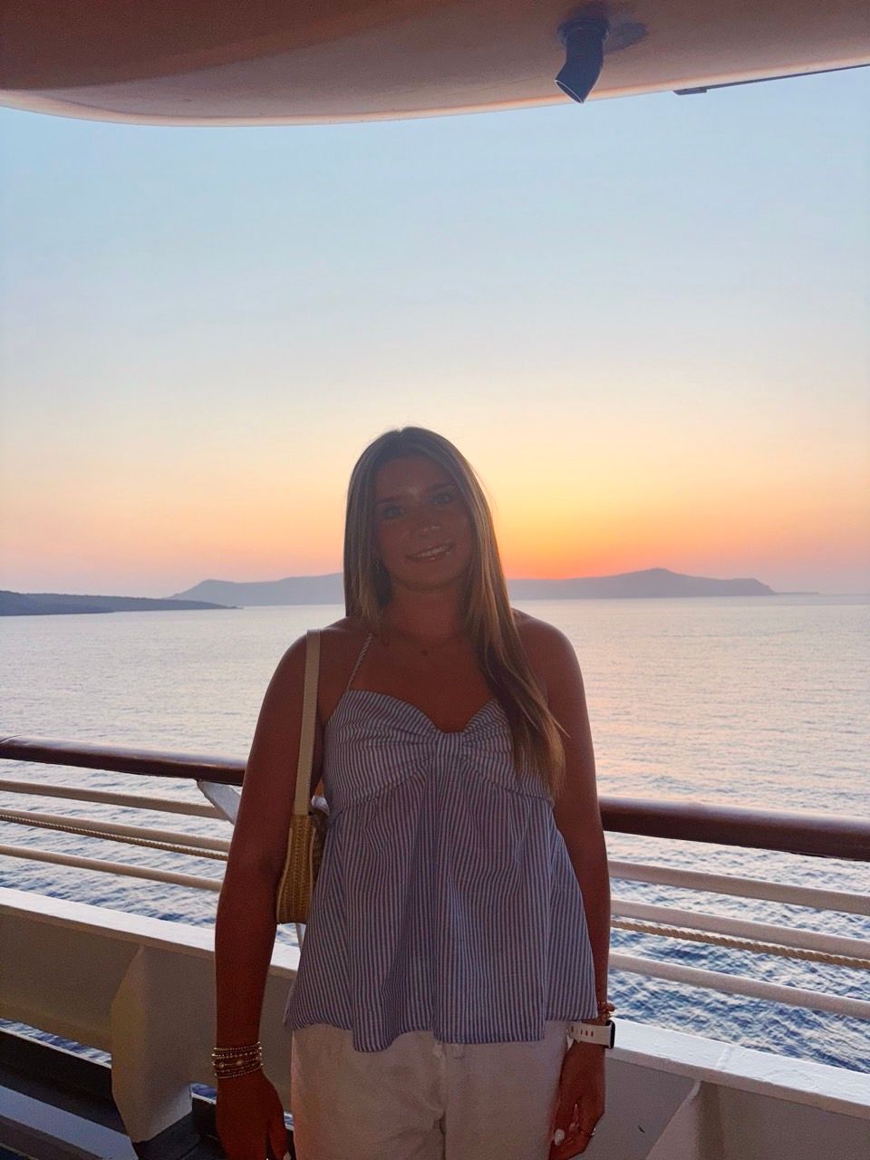 A person standing on a boat deck during sunset, with the ocean and islands in the background.