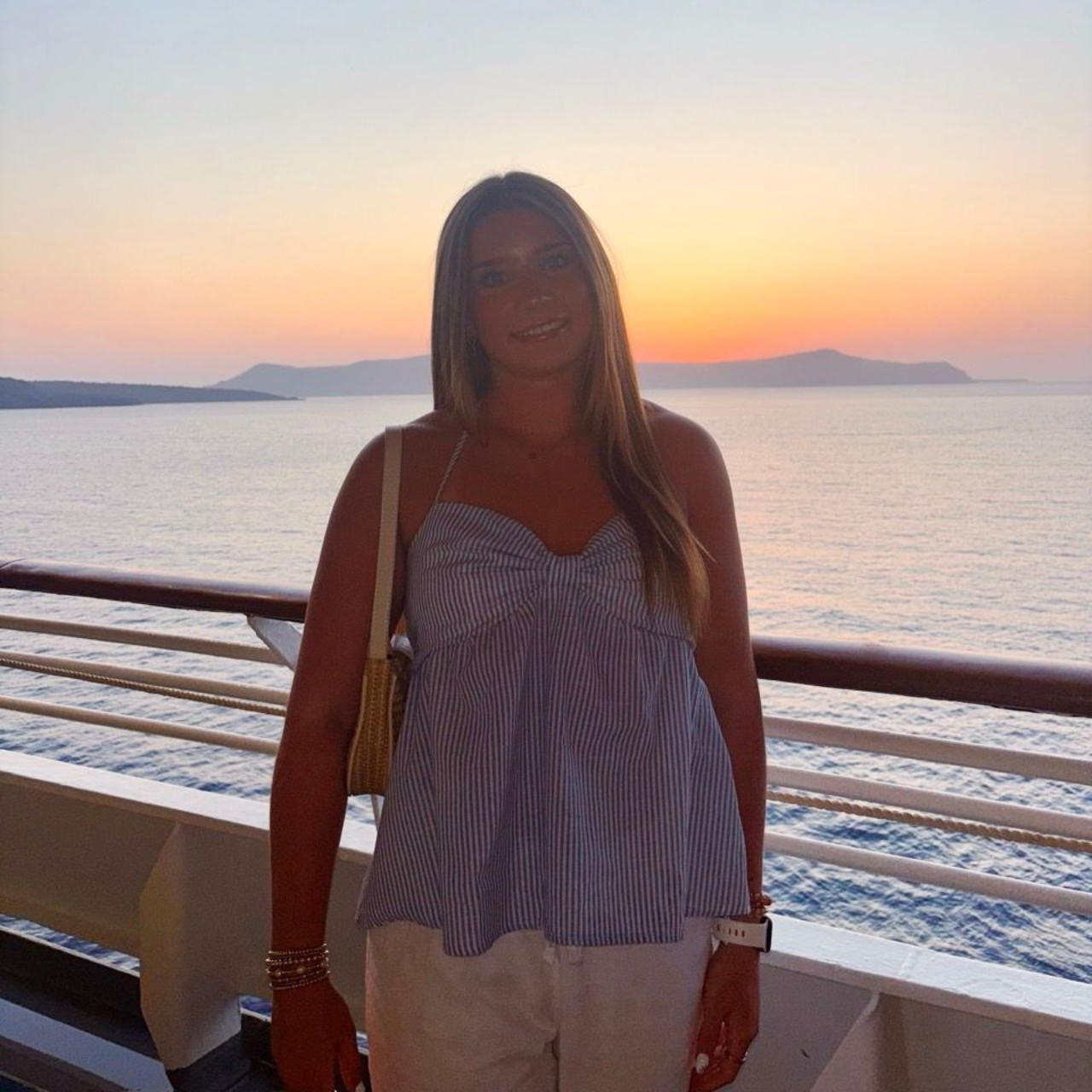 A person stands on a cruise ship deck at sunset, wearing a blue and white striped top, with the ocean in the background.