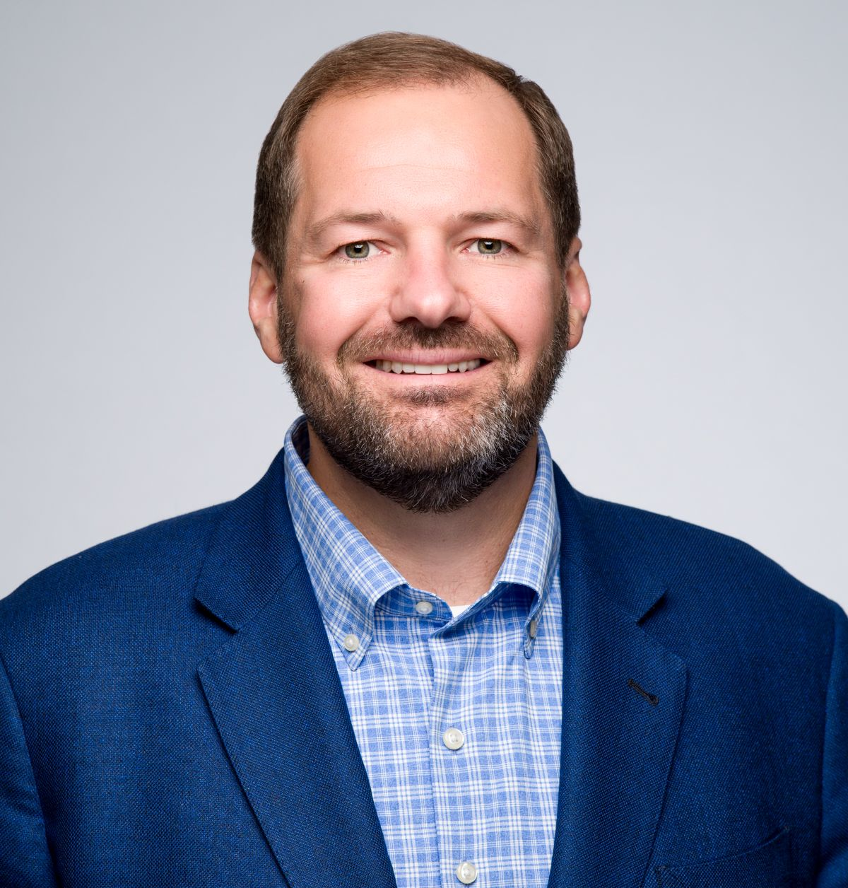 A smiling professional with a beard, wearing a blue blazer and a light blue checkered button-down shirt.