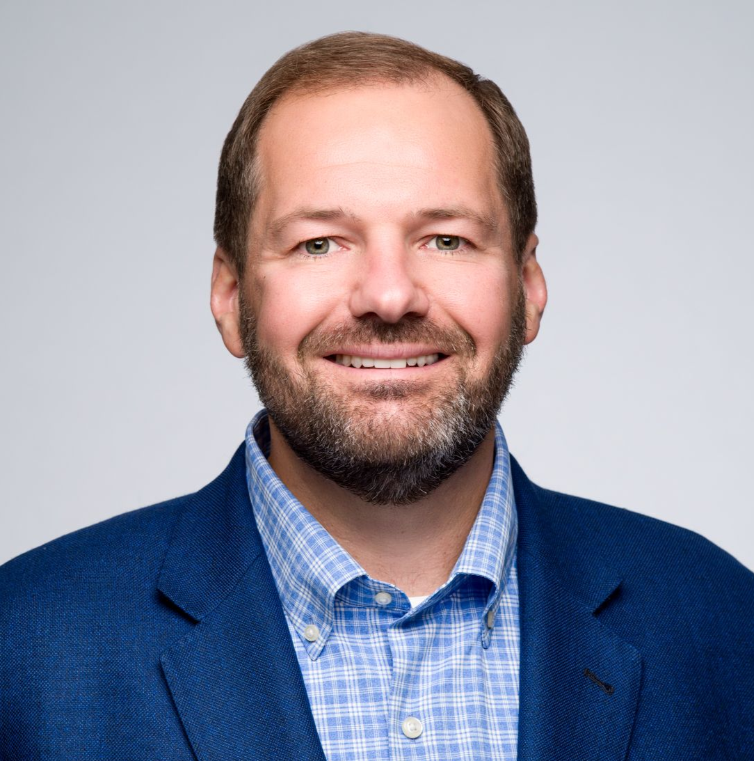 A smiling person with a beard, wearing a blue blazer and a light blue checkered button-down shirt against a grey background.