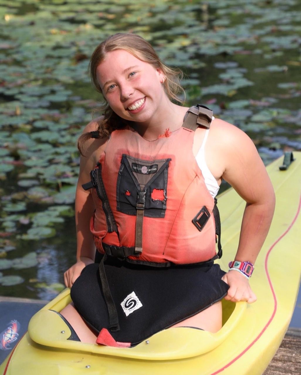 A person wearing a life vest and a spray skirt sits in a yellow kayak on a body of water with lily pads, smiling.