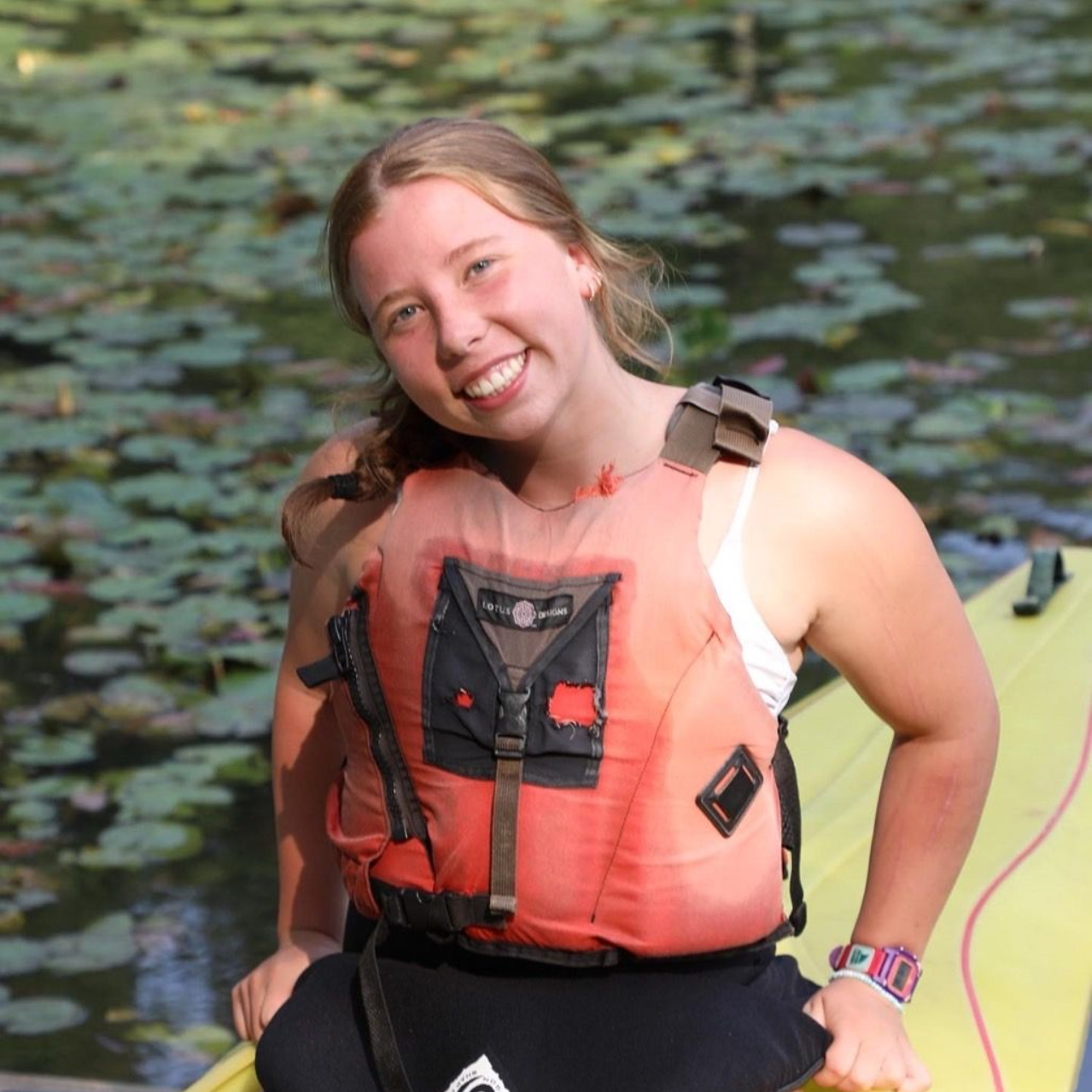 A person wearing a bright orange life vest smiles while sitting on a yellow paddleboard on a water surface with lily pads.