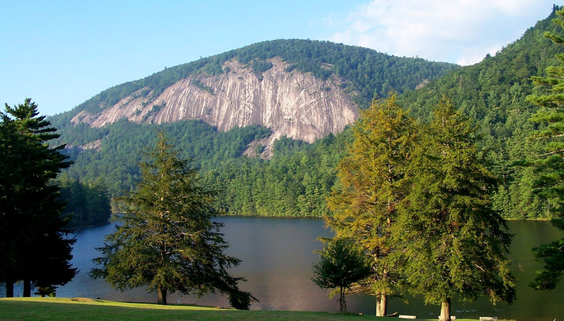 A calm lake sits beneath a prominent, steep granite mountain face surrounded by a dense, green forest under a blue sky.