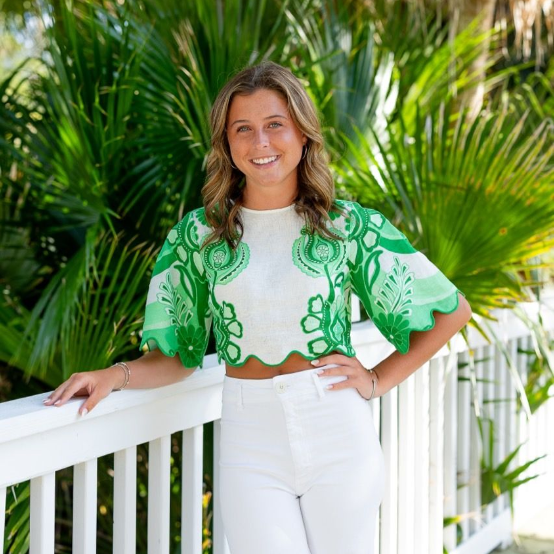 Smiling person wearing a green floral crop top and white pants, leaning on a white railing in front of palm trees.
