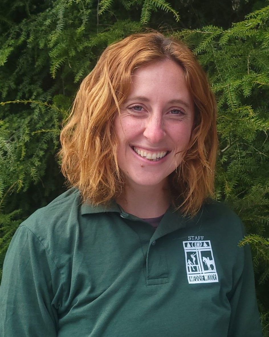 A person with shoulder-length wavy red hair smiles while wearing a dark green polo shirt in front of evergreen foliage.