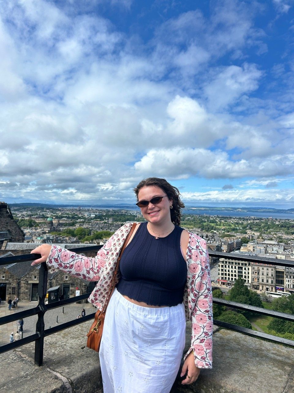 A person smiles while standing on a stone viewing platform overlooking a city skyline under a blue, cloudy sky.