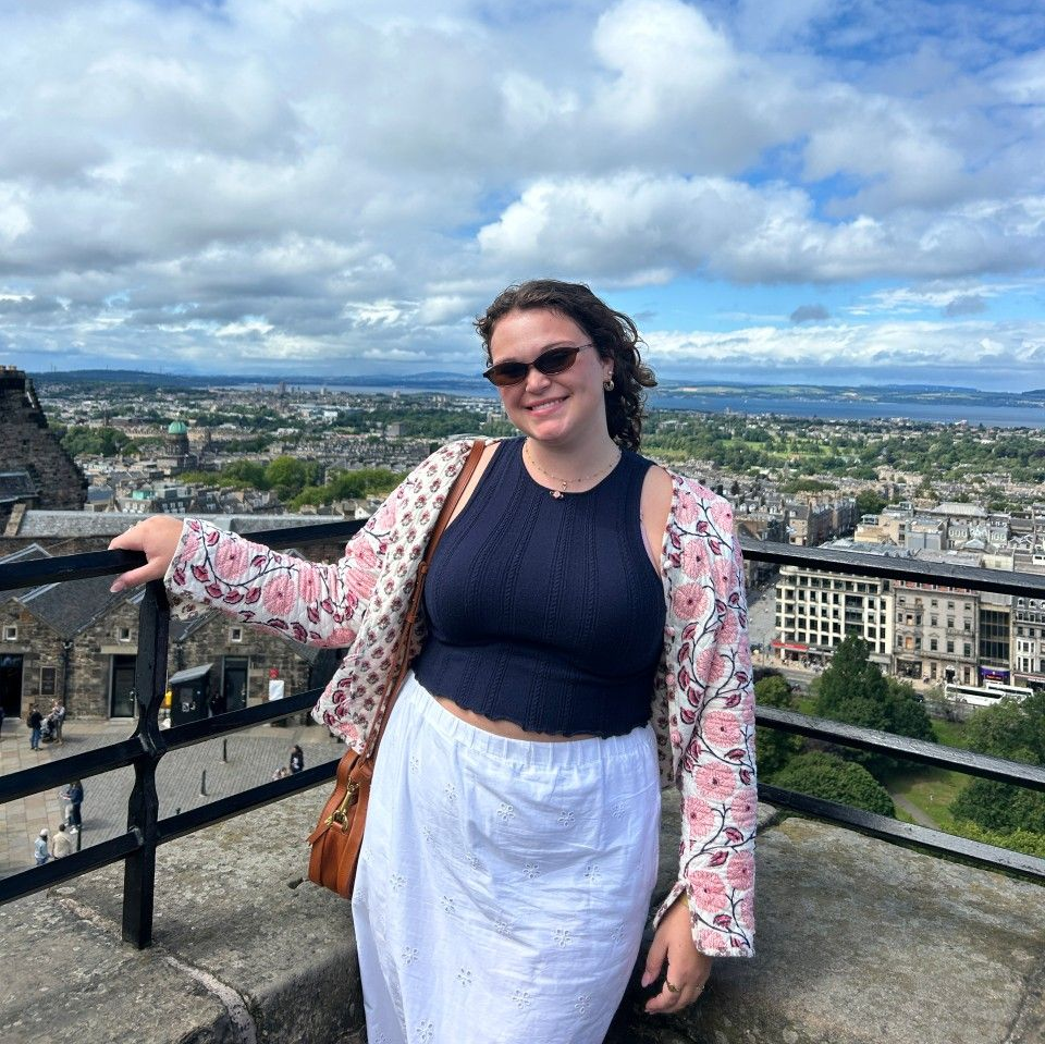 A person wearing a navy tank top and floral cardigan smiles while standing on a balcony overlooking a city and coastline.