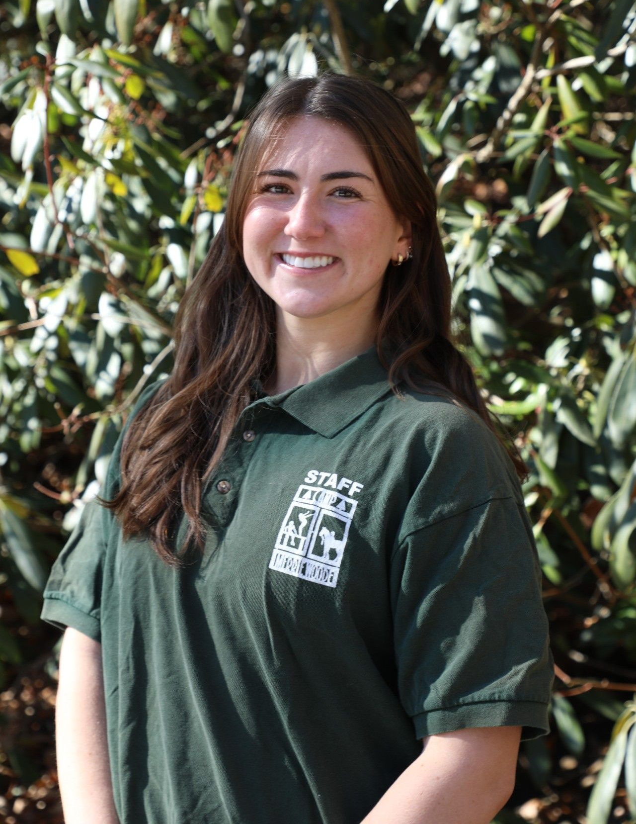 Smiling person wearing a green collared staff shirt stands outside with greenery in the background.