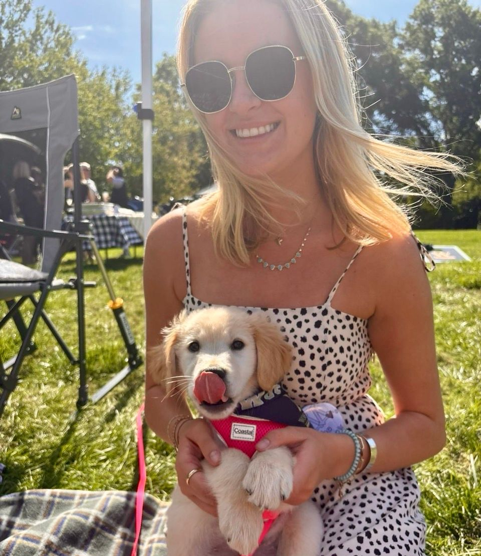 A blonde woman wearing sunglasses and a patterned dress holds a golden puppy on a sunny, grassy field.