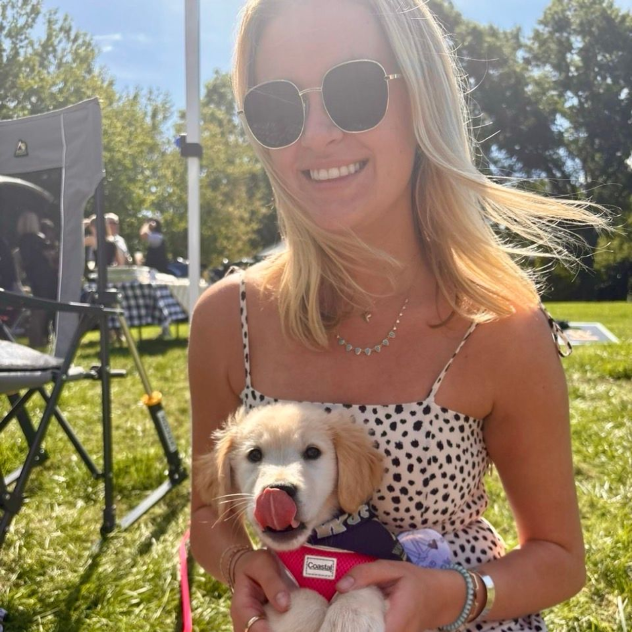 A smiling person wearing sunglasses holds a small, cream-colored puppy with its tongue out in a grassy outdoor setting.