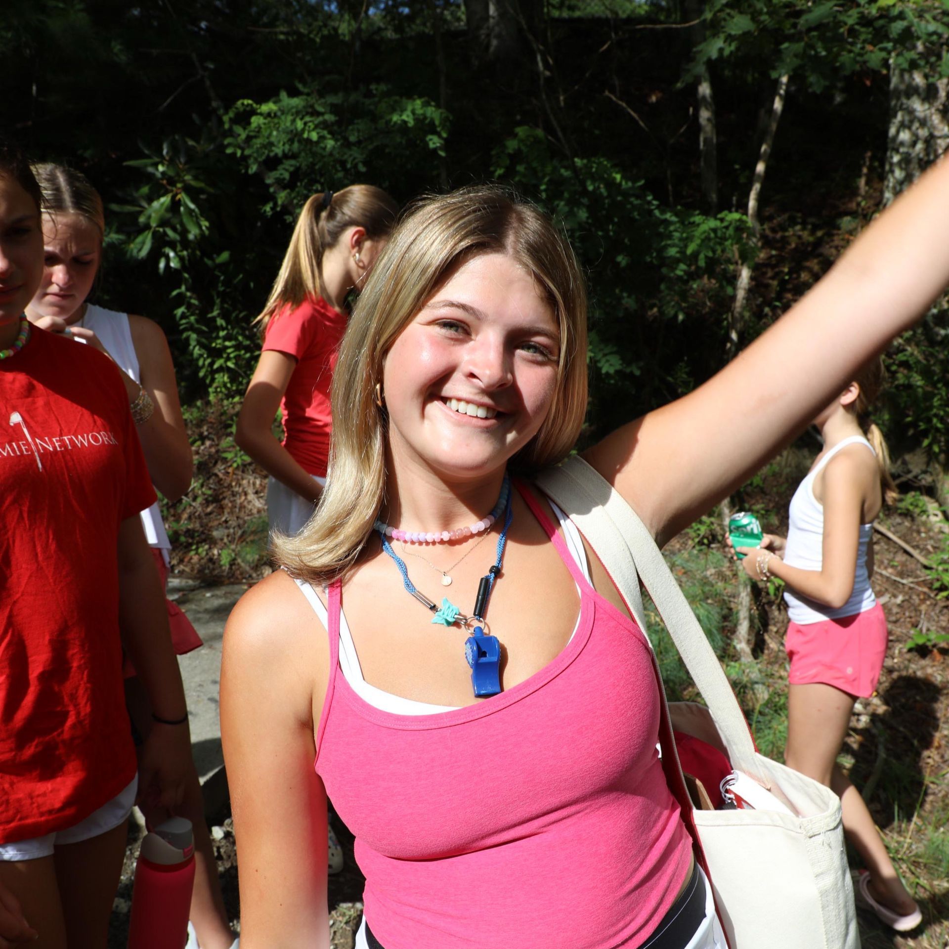 Smiling person in a pink tank top and layered necklaces stands outdoors with other people in the background.