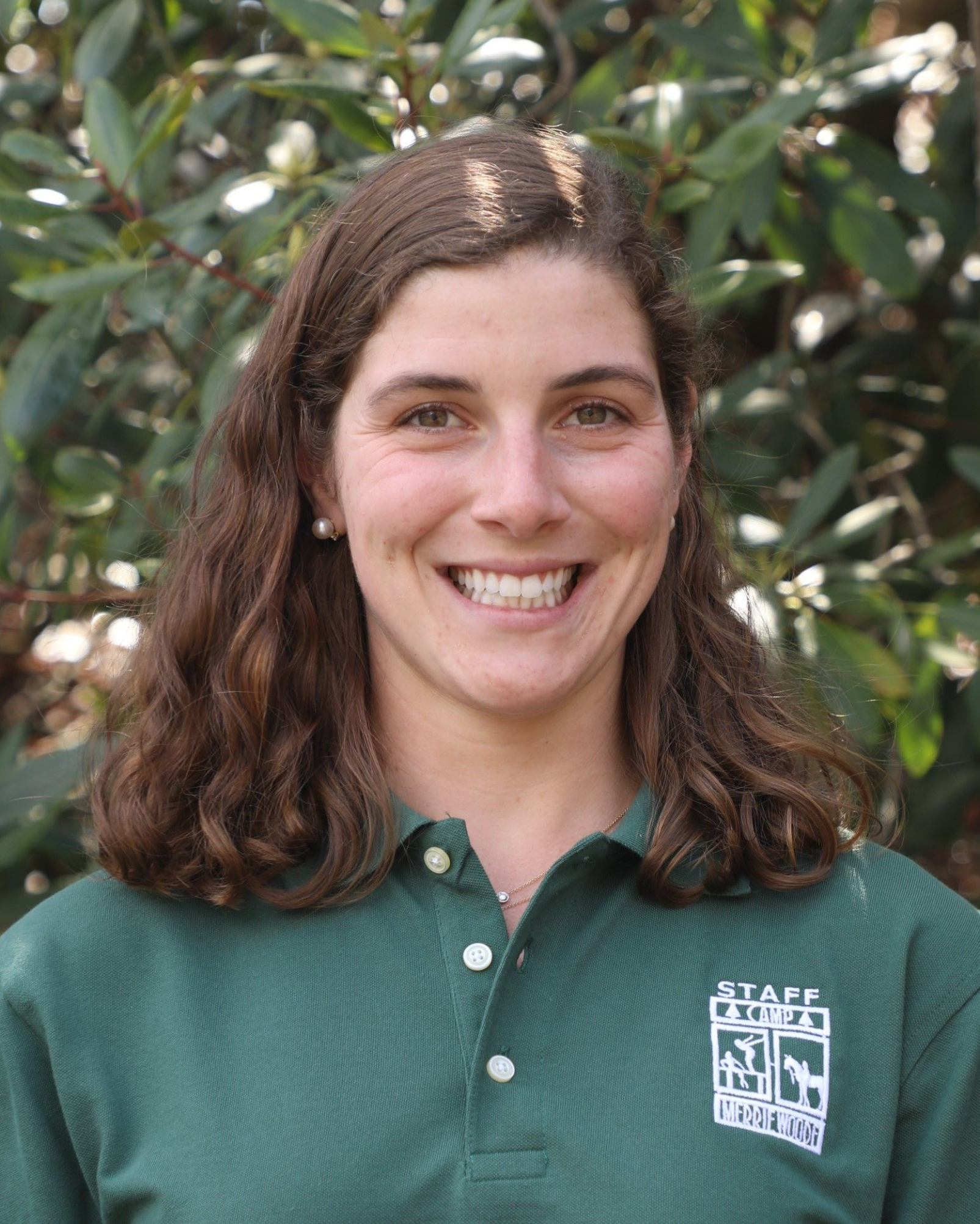 Smiling person with shoulder-length brown hair, wearing a green polo shirt with a staff logo, outdoors in front of plants.
