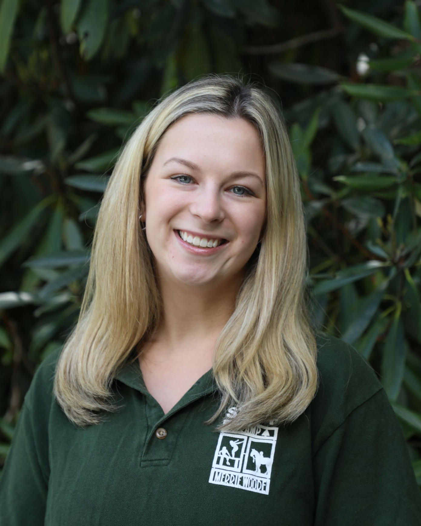 A smiling individual with long blonde hair, wearing a dark green polo shirt with a white logo, posed against leafy greenery.