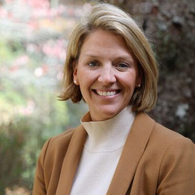 A smiling woman with blonde hair, wearing a beige turtleneck and brown blazer, standing in a blurred, wooded setting.