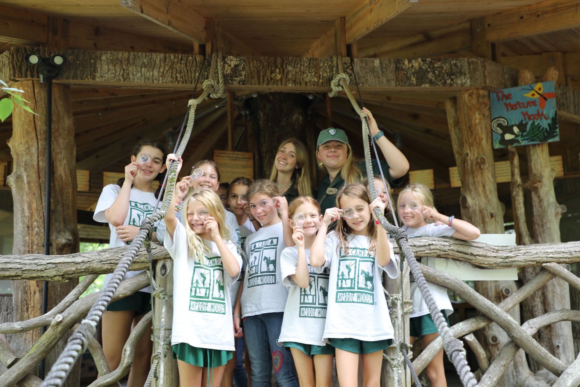 A group of people wearing matching white t-shirts pose on a rustic, wooden treehouse platform.