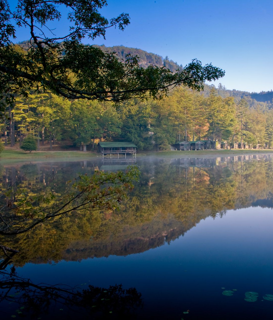A calm, misty lake reflects a dense forest and a distant mountainside under a clear, bright blue morning sky.
