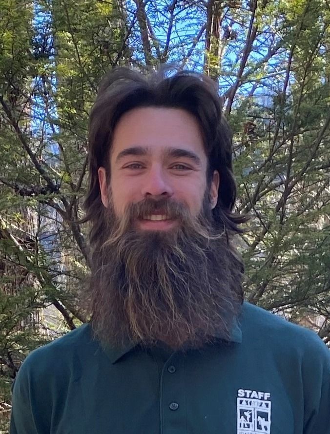 A person with long, dark hair and a full beard wearing a green polo shirt with a staff logo, posed in front of trees.