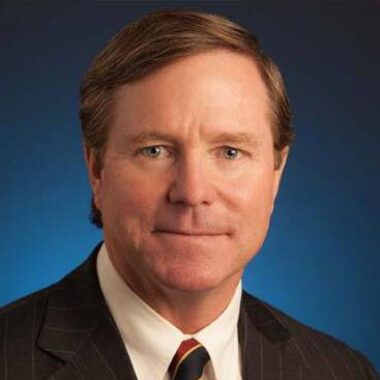 Headshot of a person in a pinstripe suit, white shirt, and patterned tie against a blue background.