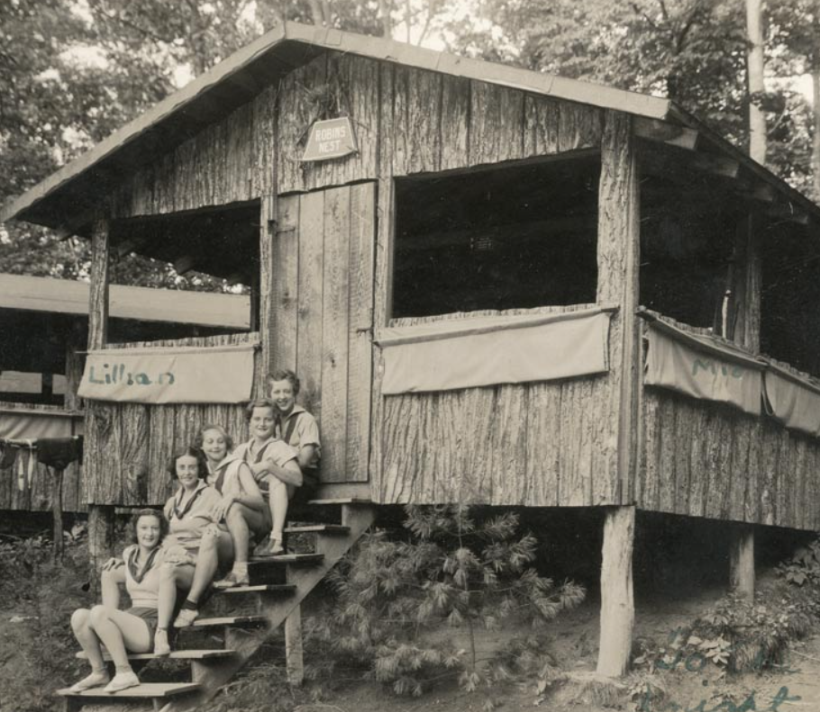 Five people sit on the steps of a rustic, bark-covered wooden cabin in a forest clearing.