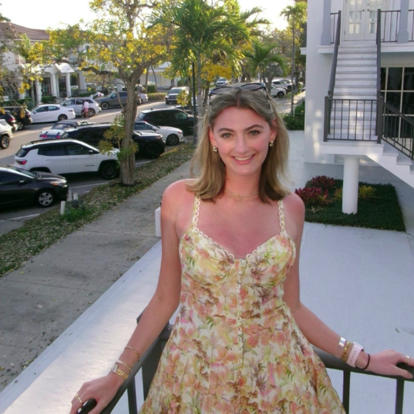 A person smiles for a photo outdoors, wearing a pink tank top and a blue whistle necklace, with others in the background.