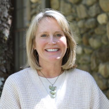 A smiling person with shoulder-length blonde hair wearing a white textured sweater and a leaf-shaped pendant necklace.