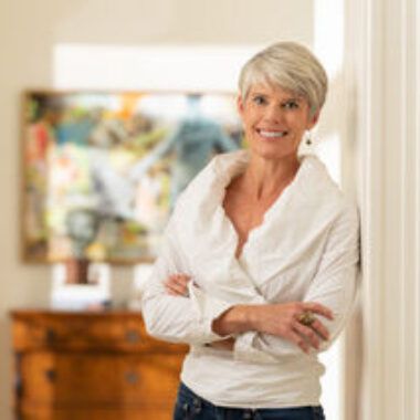 A person with short, light-colored hair, wearing a white, long-sleeved top with arms crossed, stands indoors near art.
