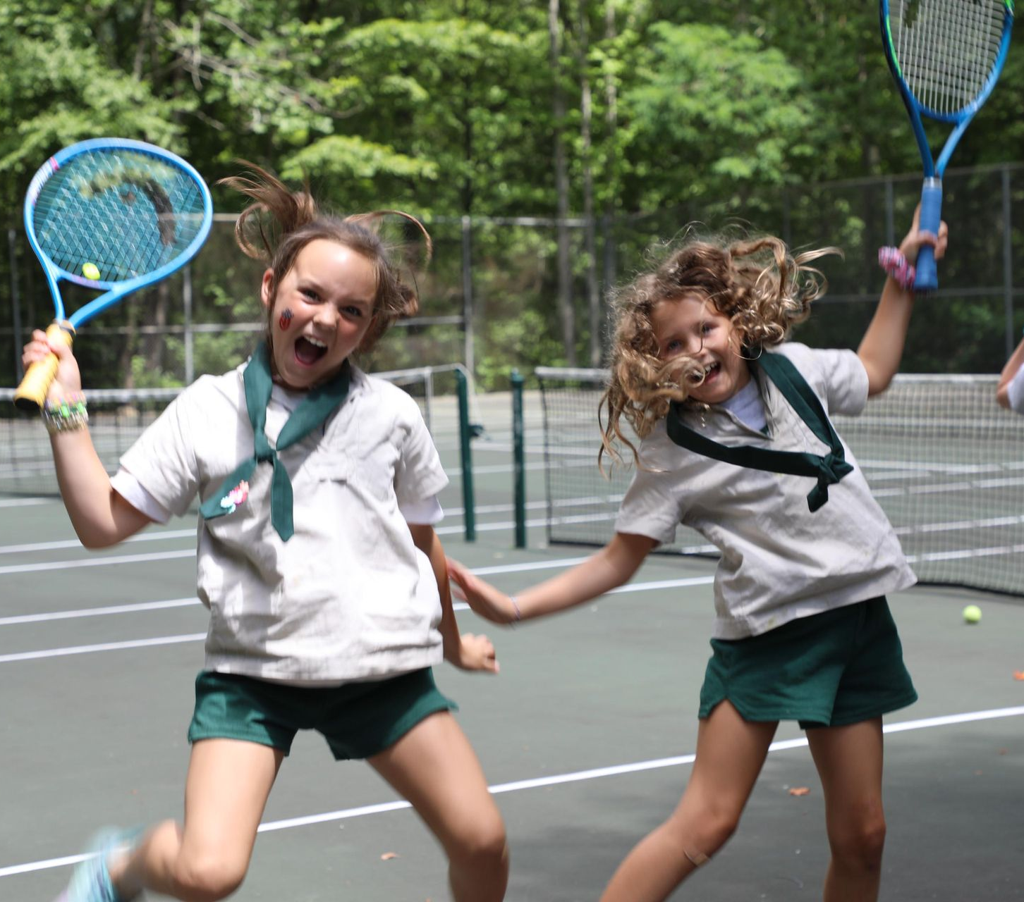 Two people in white shirts and green shorts jump on a tennis court, holding rackets and smiling with joy.