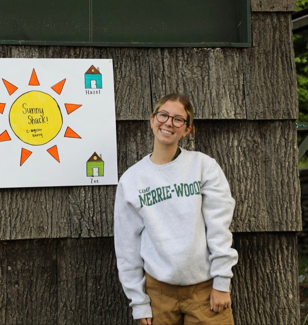 A person in a Merrie-Woode sweatshirt smiles beside a sign with a sun drawing and two small house illustrations.