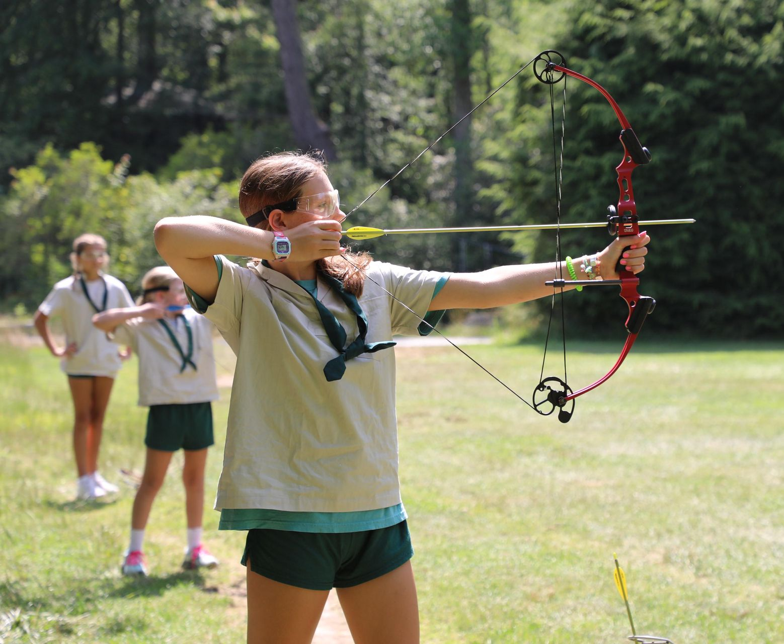 A person in a camp uniform aims a red compound bow at a target in a grassy field, with two others standing in the background.