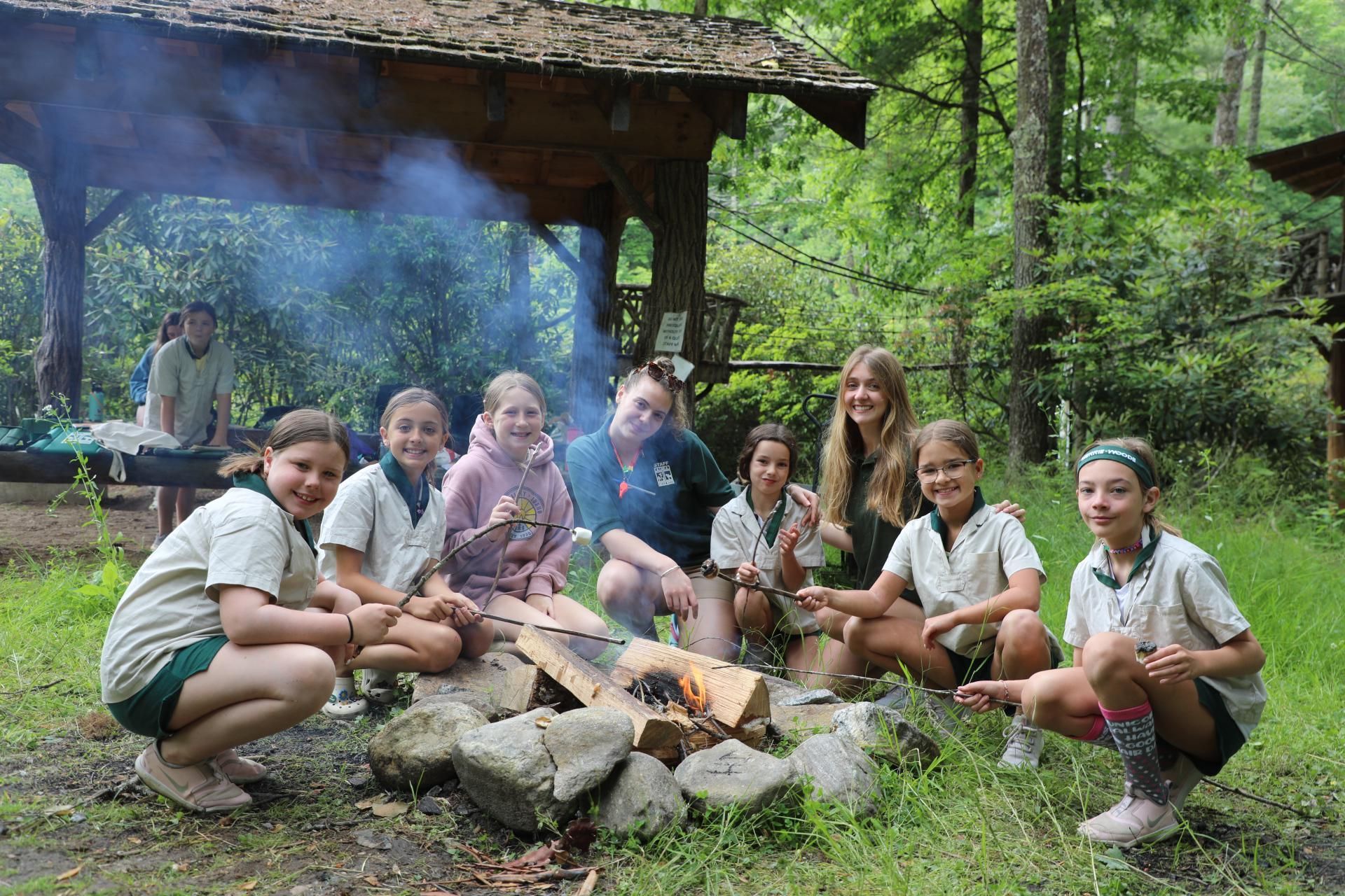 A group of young people in camp uniforms sit around a small campfire in a wooded area near an open wooden shelter.