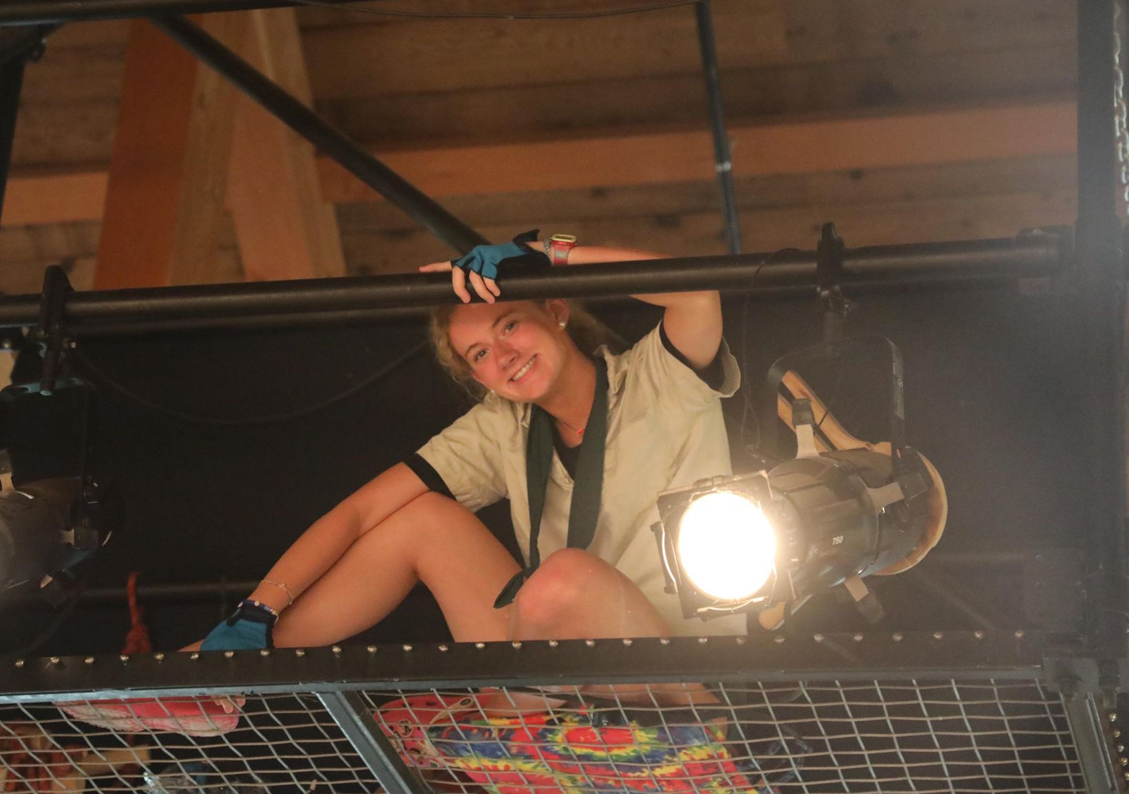 A smiling person sits on a metal catwalk above a stage, holding a lighting truss next to a bright stage spotlight.