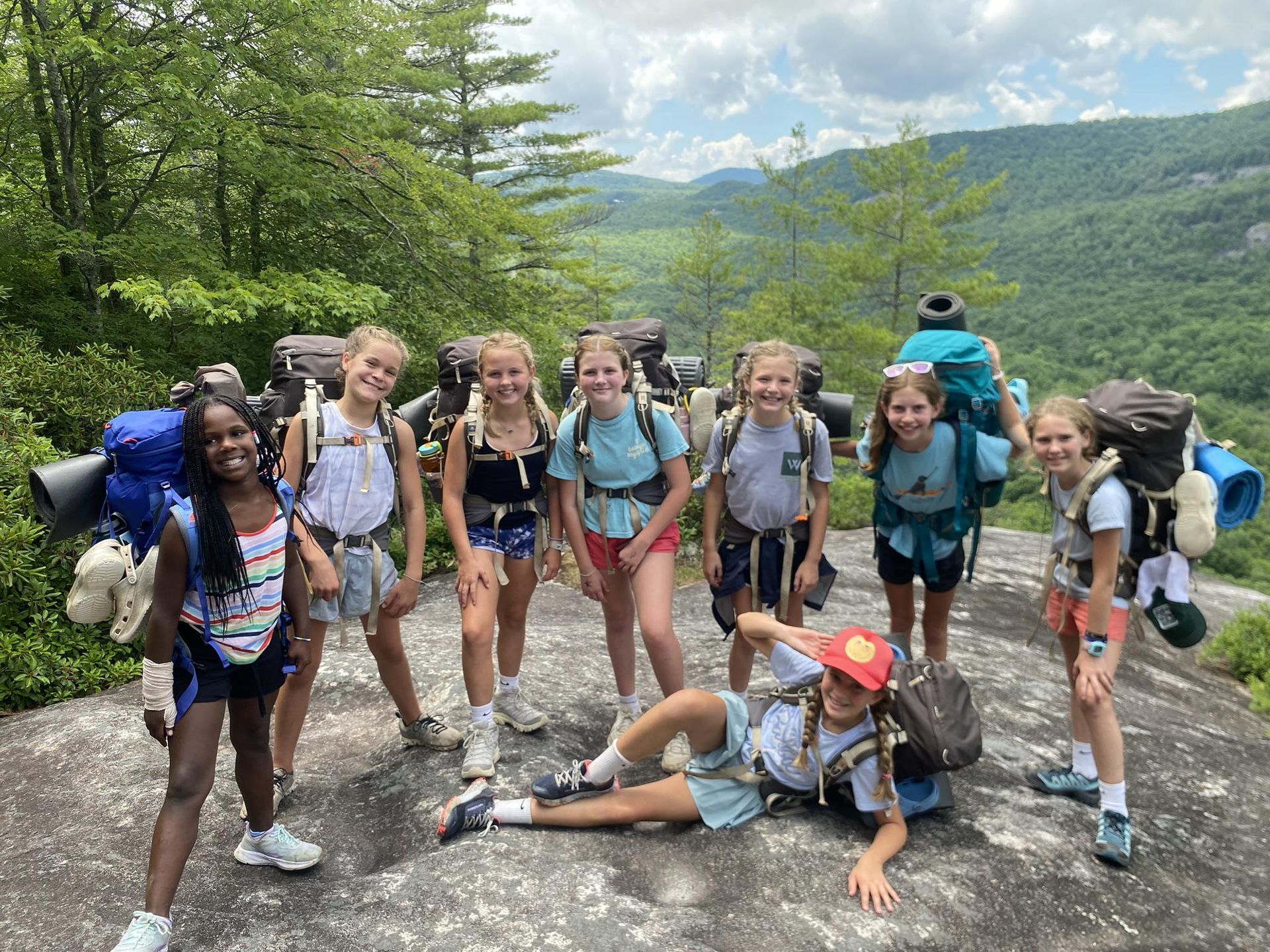 A group of hikers with large backpacks smile on a rocky mountain overlook with green forested hills in the background.