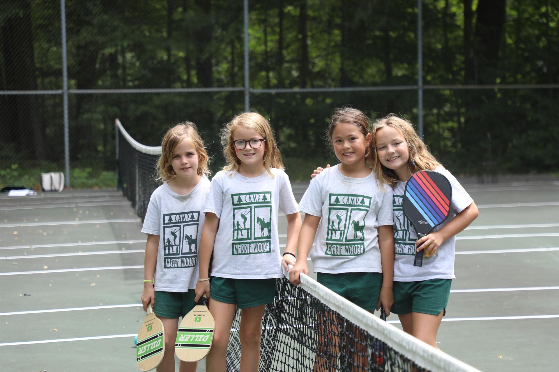Four children in matching white shirts and dark shorts stand smiling behind a pickleball net on an outdoor court.