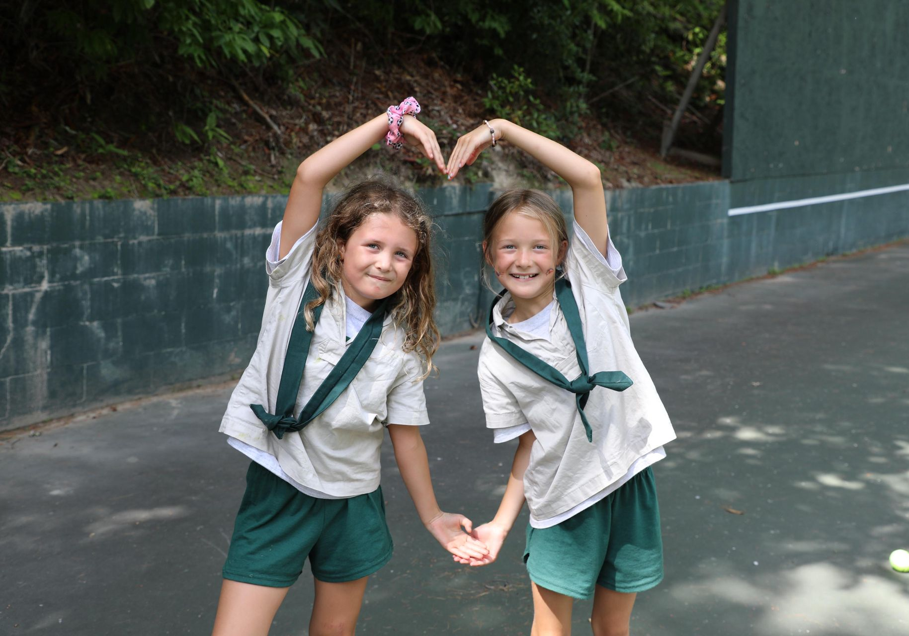 Two children in khaki uniform shirts and green shorts smiling and forming a heart shape with their arms outdoors.