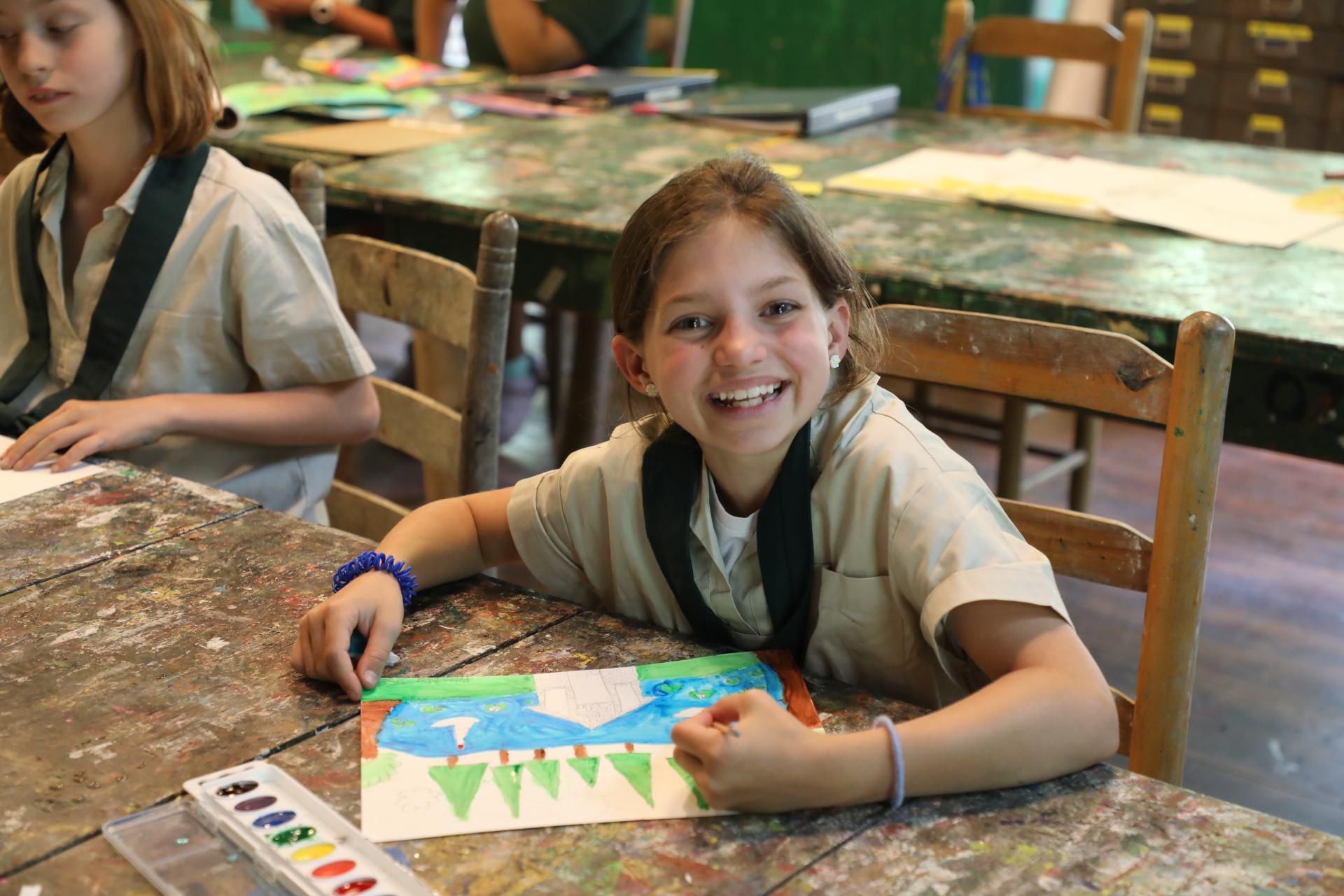 A smiling child sits at a worn wooden table in an art room, holding a marker while coloring a landscape drawing.
