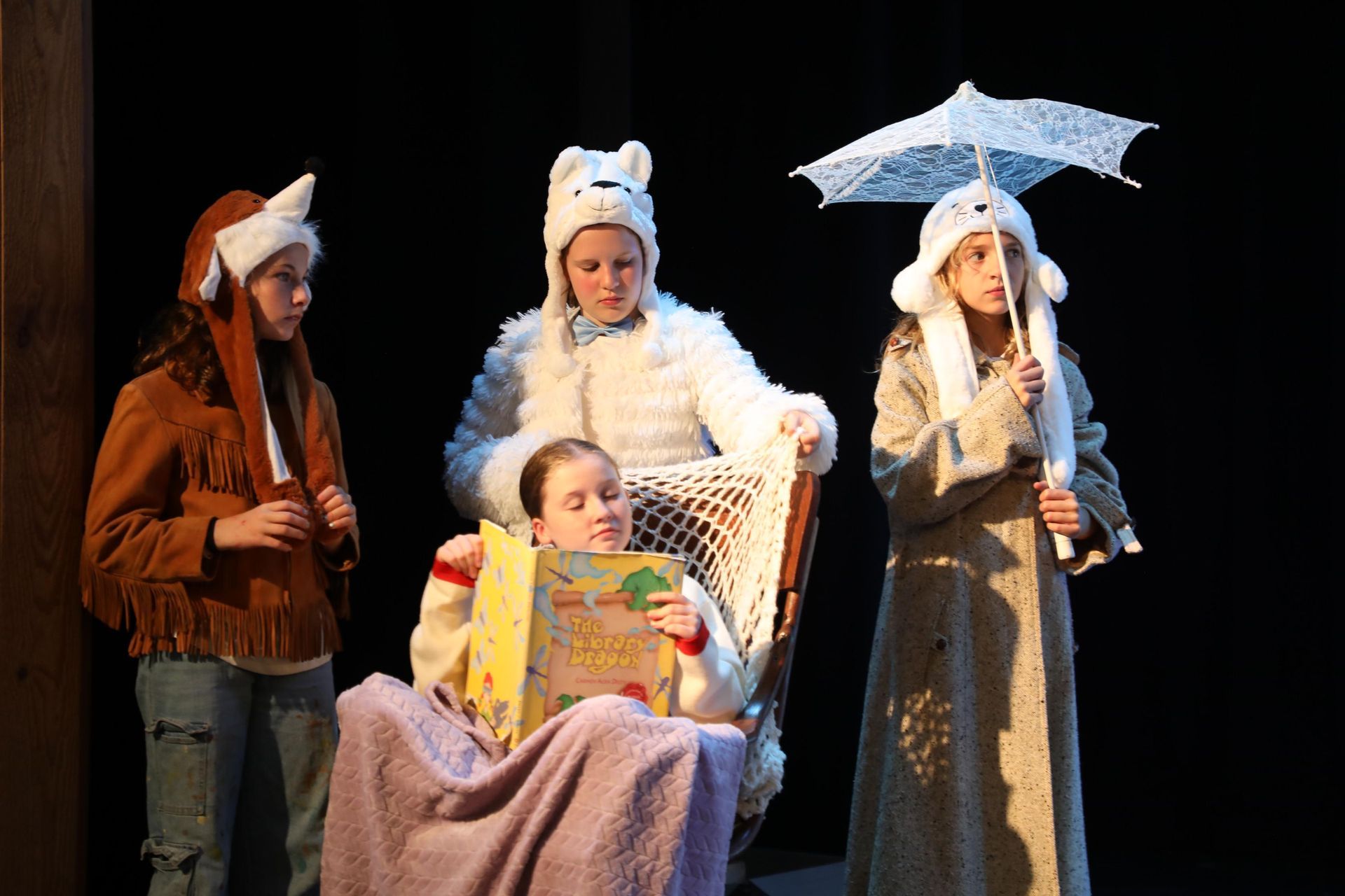 Four people in costumes stand and sit on a stage, one reading a book, while another holds a white parasol.