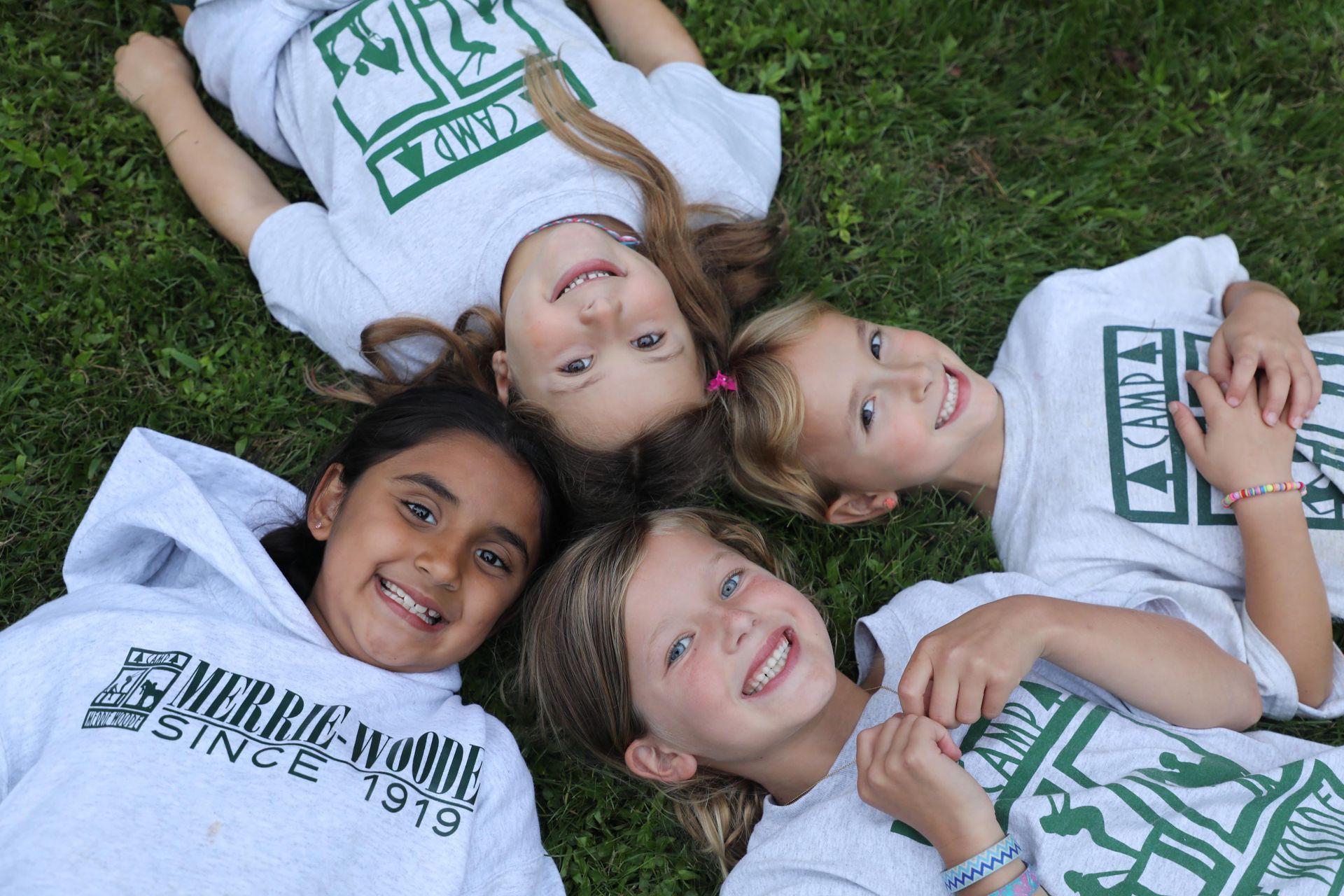 Four children lie on grass in a circle, heads together, wearing matching camp T-shirts and smiling at the camera.