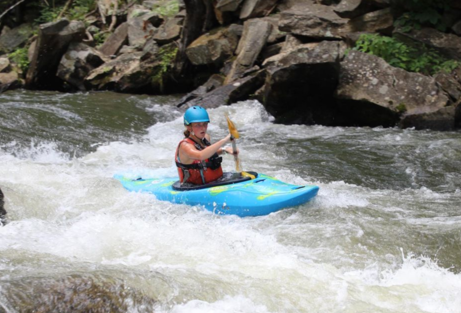 A person wearing a helmet and life vest paddles a bright blue kayak through rushing river rapids near rocky banks.