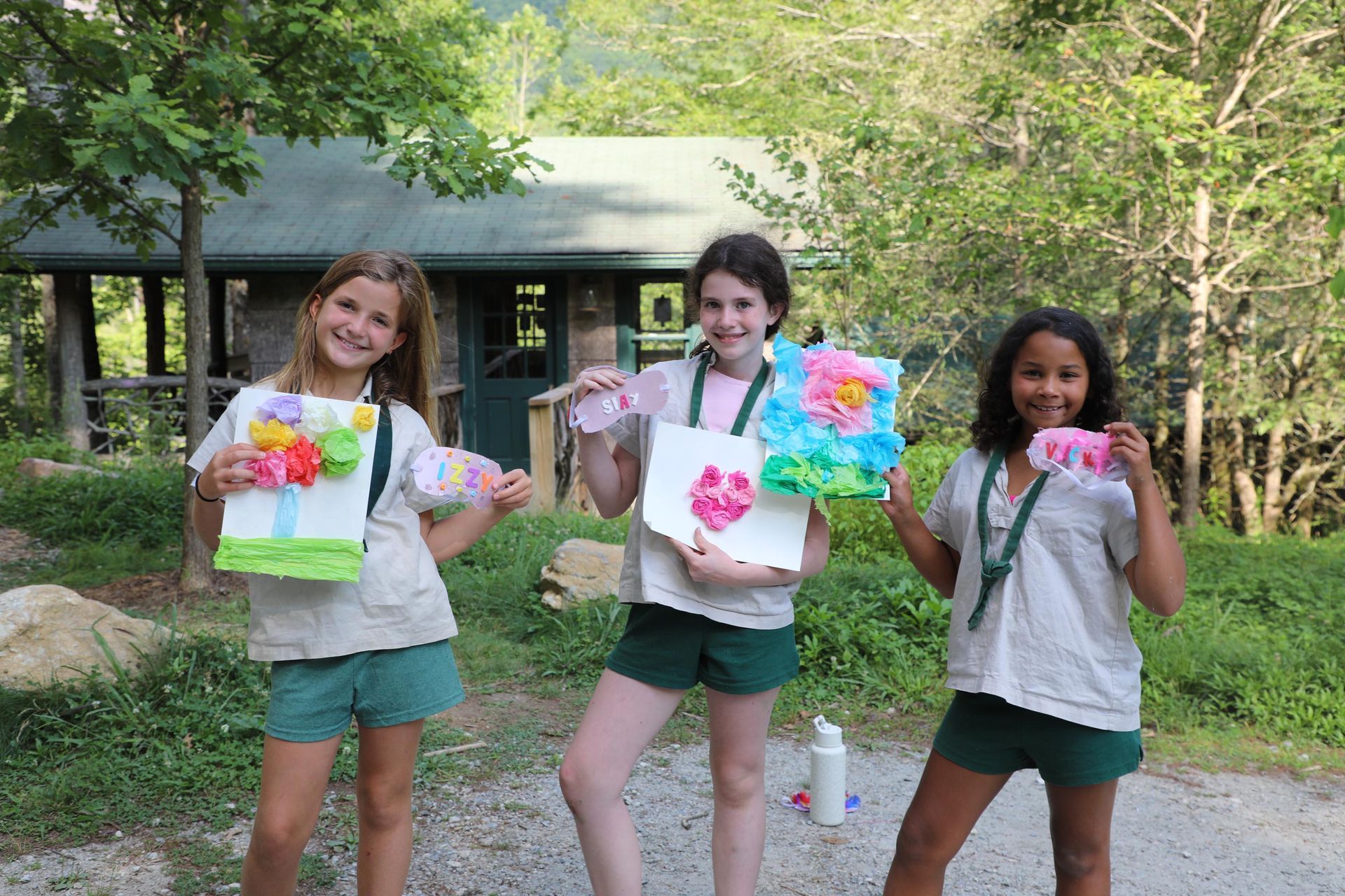 Three children in camp uniforms stand outdoors holding colorful paper crafts in front of a rustic cabin.