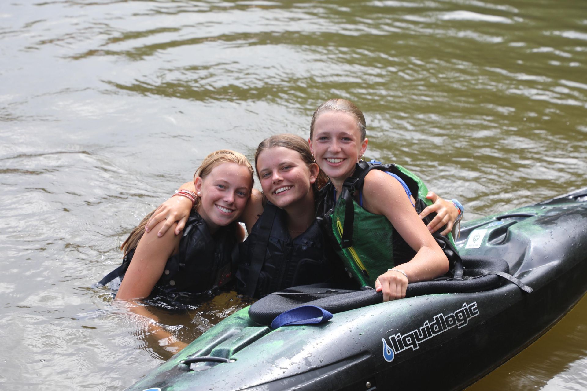 Three people in life jackets smile while partially submerged in water next to a black kayak.