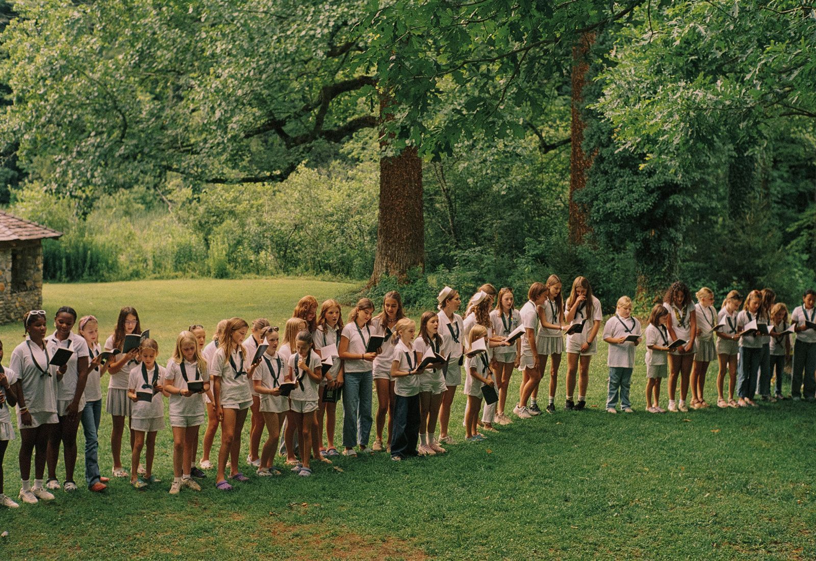 A large group of people in white attire sits on stone steps and grass, gathered for an outdoor event under trees.