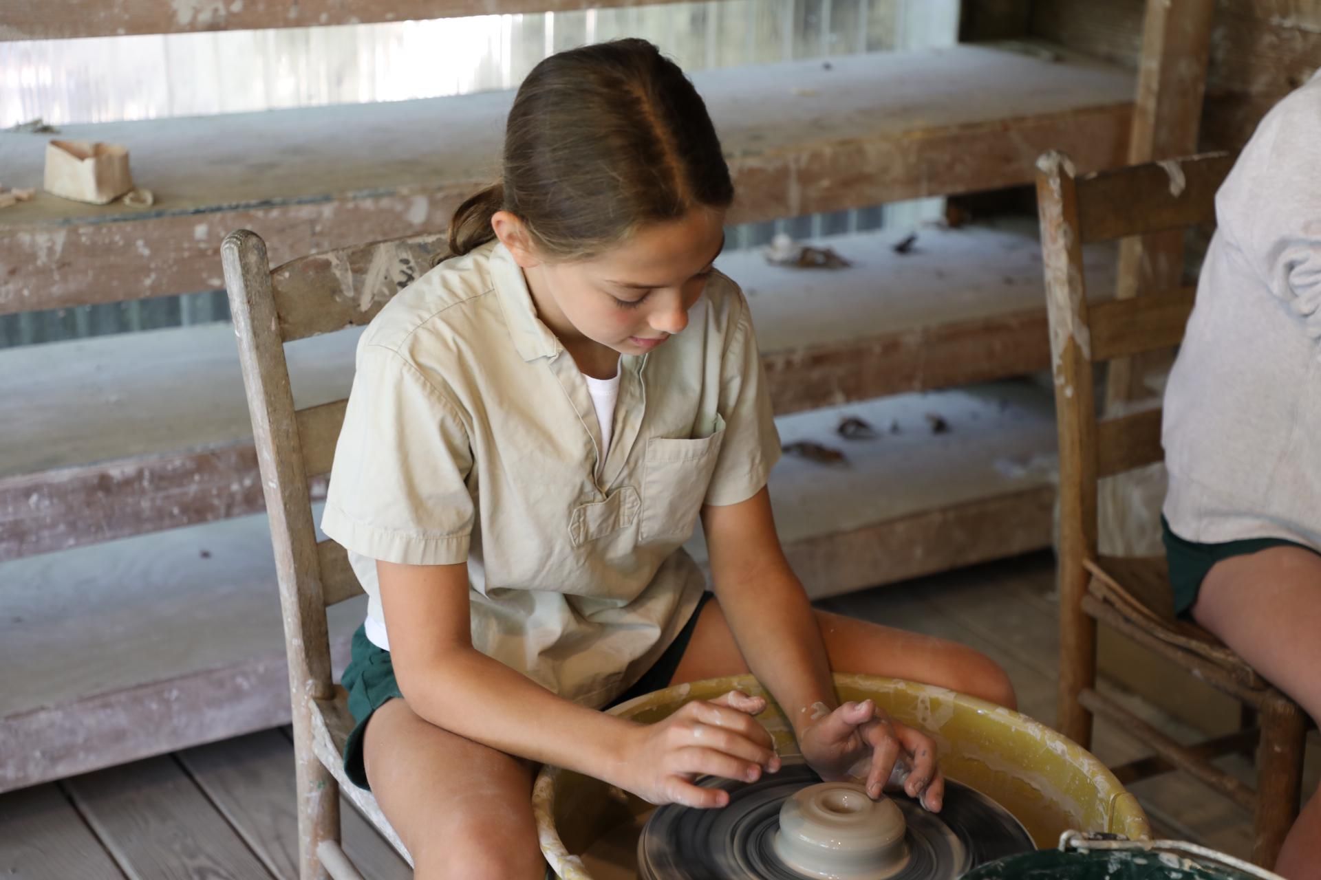 A person in a beige shirt uses a pottery wheel to shape a clay bowl in a rustic, workshop-style studio.