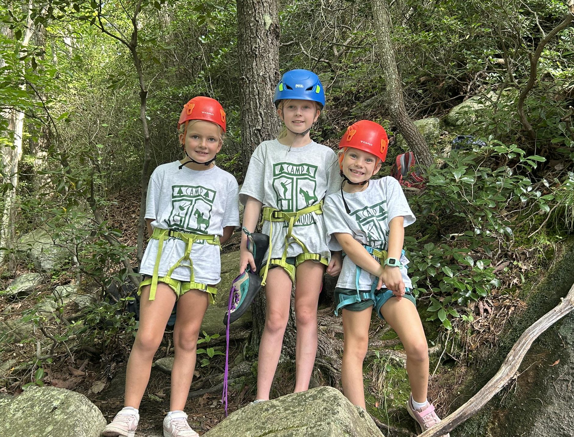 Three children in matching t-shirts and helmets stand on a rocky, wooded path while wearing climbing harnesses.