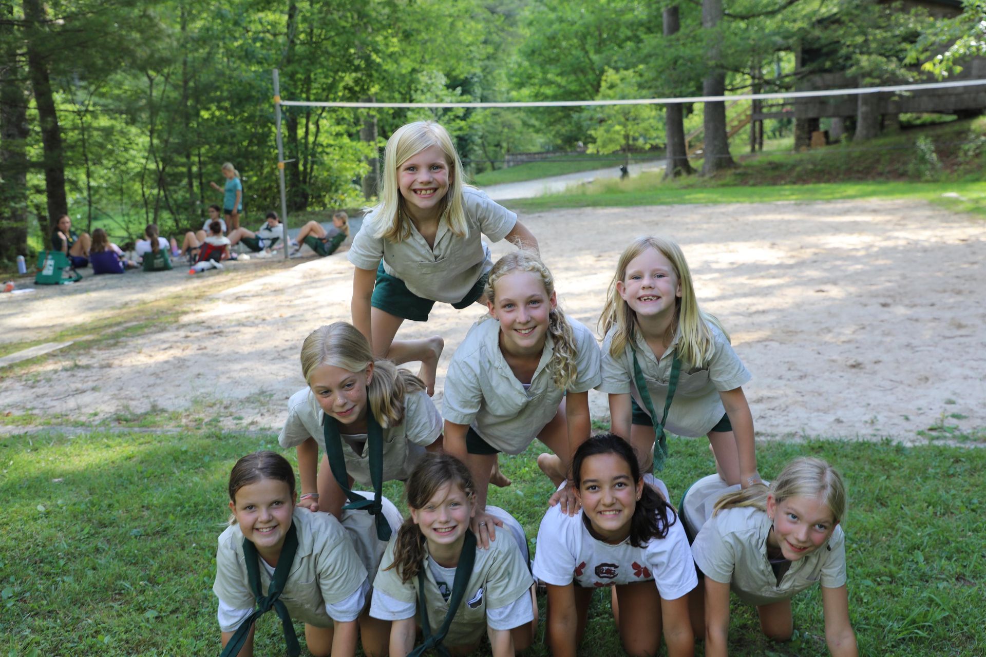 A group of smiling people form a human pyramid on a grassy field in front of a sand volleyball court.