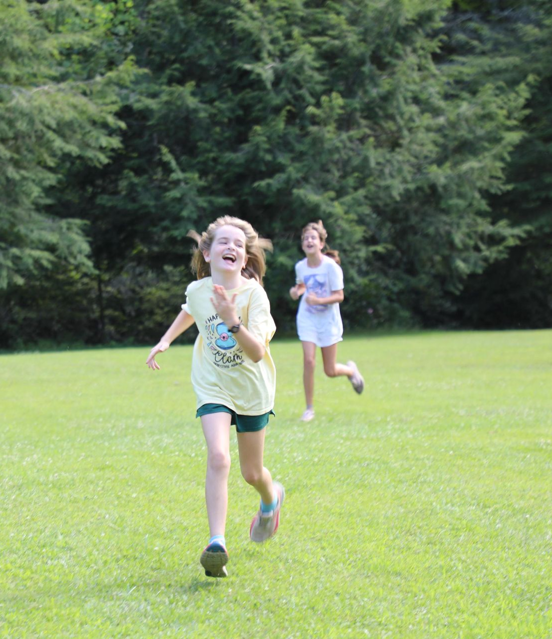 Two children running across a sunny, green grassy field in front of a line of trees, both laughing.