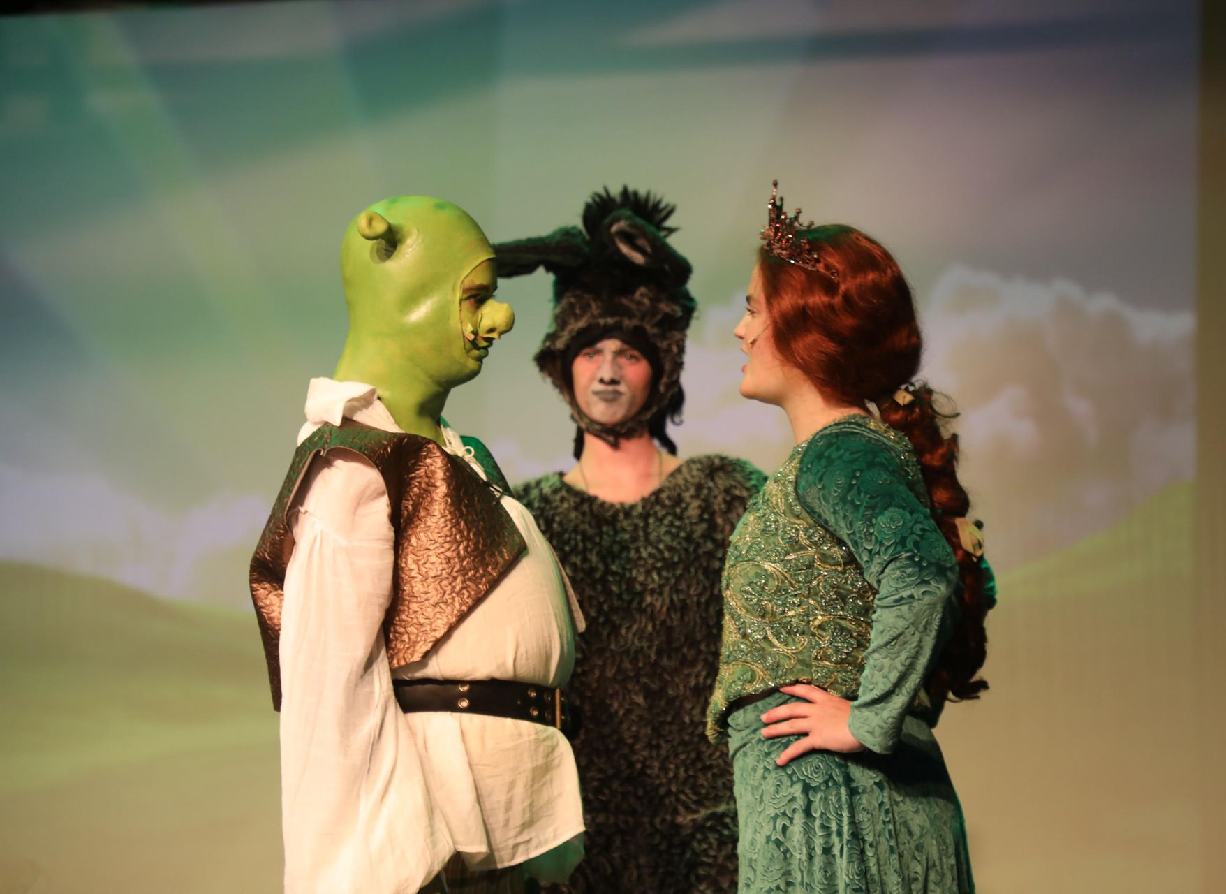 Costumed actors portraying Shrek, Donkey, and Fiona in a stage production, standing together against a painted backdrop.