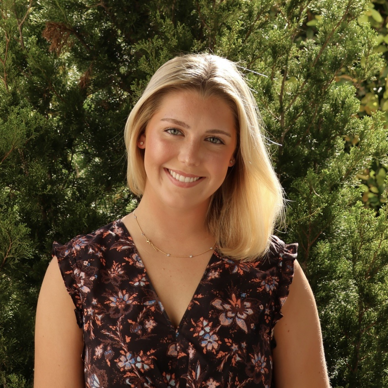 A person wearing a navy tank top and floral cardigan smiles while standing on a balcony overlooking a city and coastline.