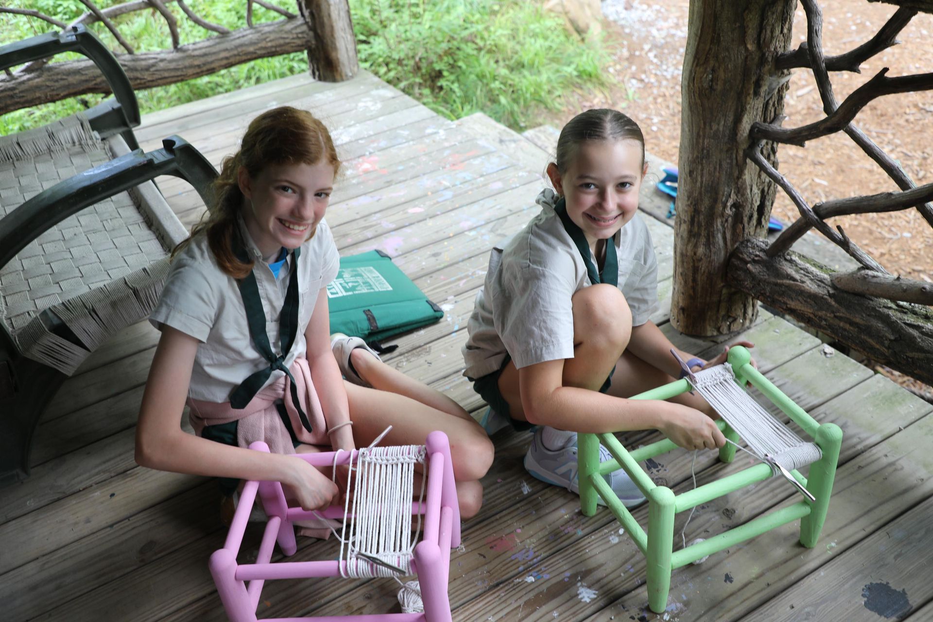 Two people sit on a wooden deck weaving yarn on small, colorful looms in an outdoor, shaded setting.