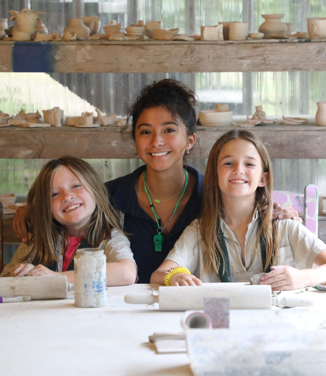Three people smiling behind a workbench covered in clay tools and rolling pins, with shelves of pottery in the background.