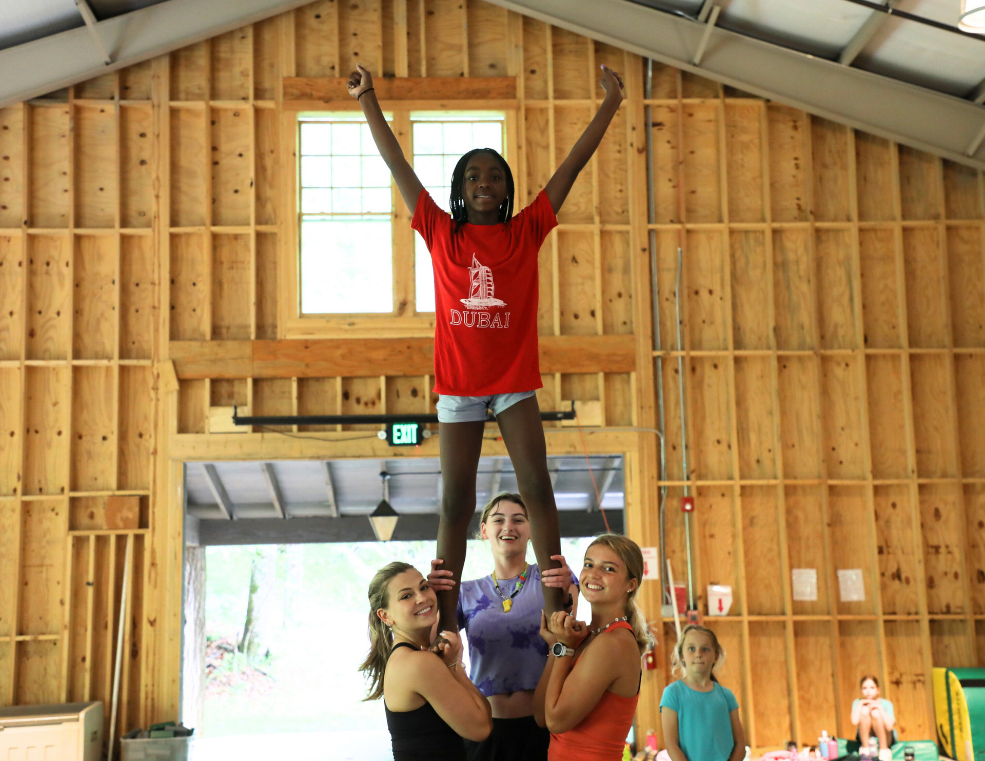 A person in a red shirt stands on the shoulders of two people, arms raised, in front of a wooden-framed structure.